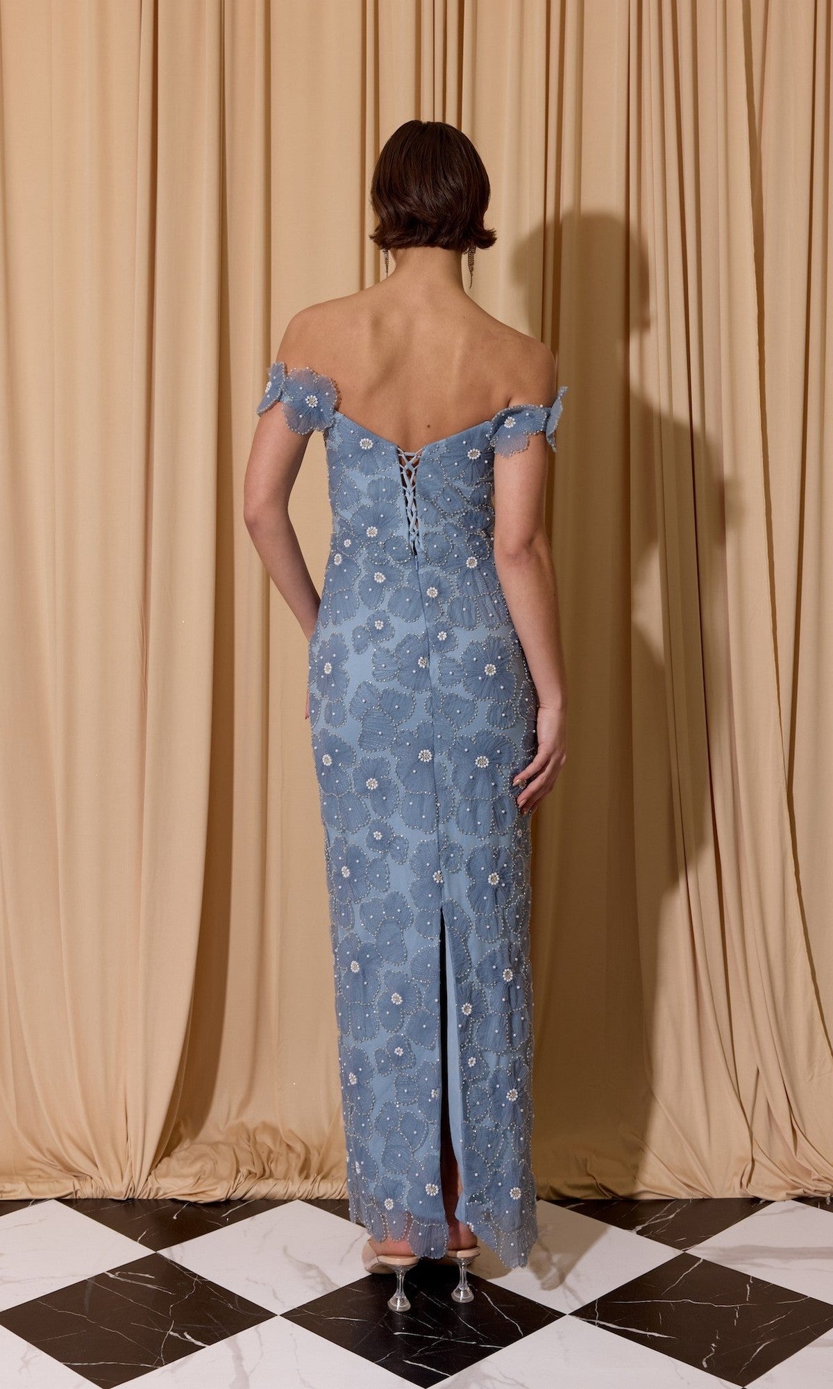 A person stands on a black and white tile floor, facing beige curtains, wearing the Eureka EK205 long prom dress in light blue floral lace with off-the-shoulder sleeves, a lace-up back, and a slit at the hem.